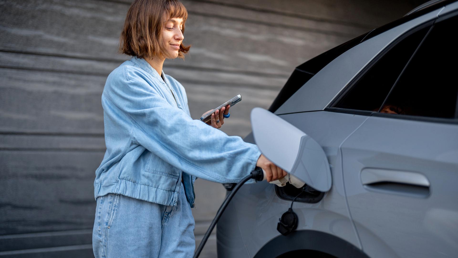 En ung kvinna med kortklippt hår laddar sin elbil samtidigt som hon kollar på sin mobiltelefon.