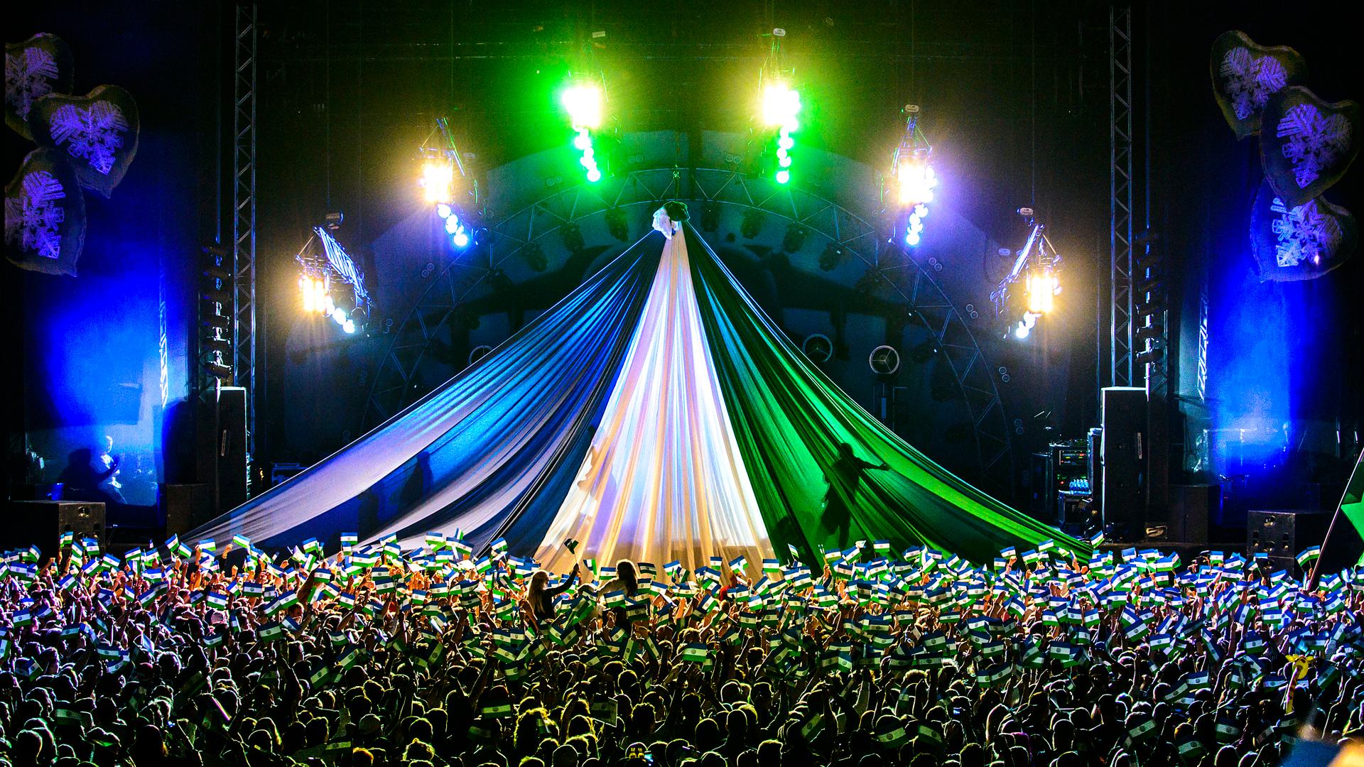 Presidenttalet på scen under Storsjöyran. Ett stort skynke i blått, vitt och grönt hänger över en hel scen och flera lampor lyser i samma färger. Framför scenen syns ett folkhav där många människor håller upp Jämtlandsflaggan.