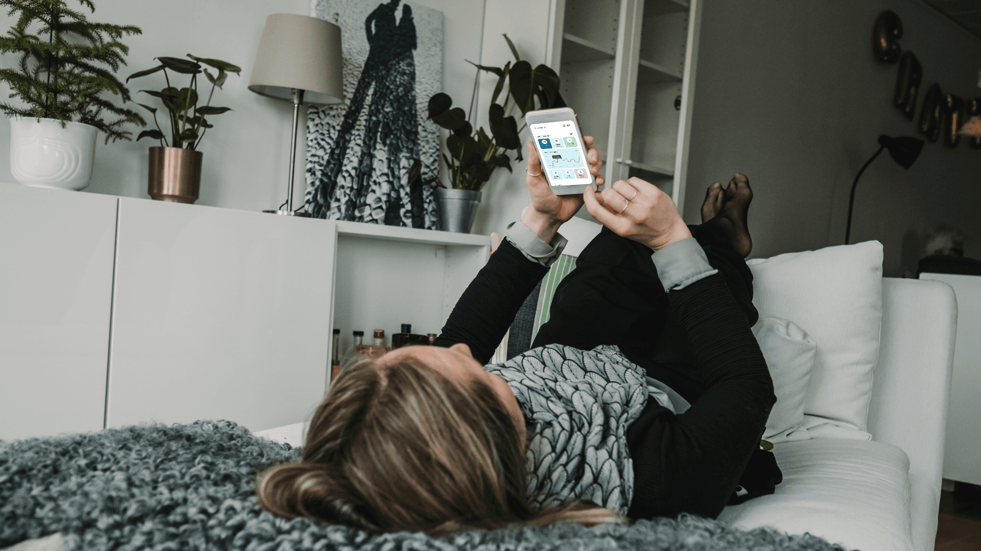 En kvinna ligger på rygg i soffan och scrollar på sin telefon.