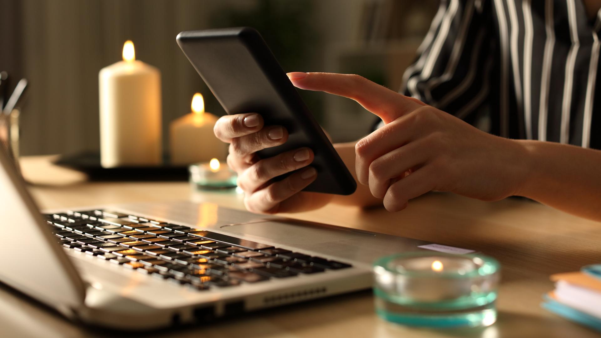 Närbild på en persons händer som håller i en telefon. Personen sitter vid ett skrivbord och i bild syns flera levande ljus och en dator.