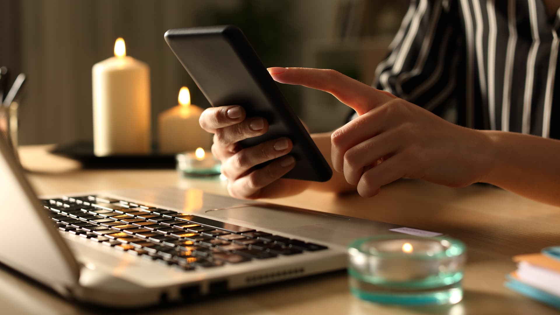 Närbild på en persons händer som håller i en telefon. Personen sitter vid ett skrivbord och i bild syns flera levande ljus och en dator.