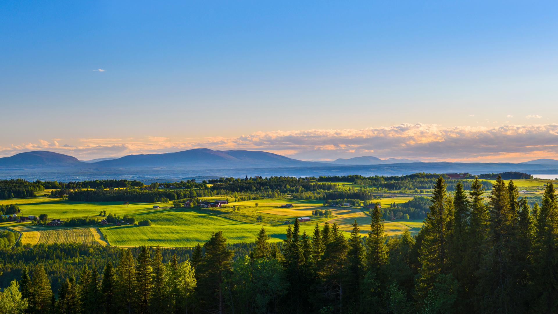 Solnedgång över ett jämtländskt landskap med skog, åkrar och fjäll.