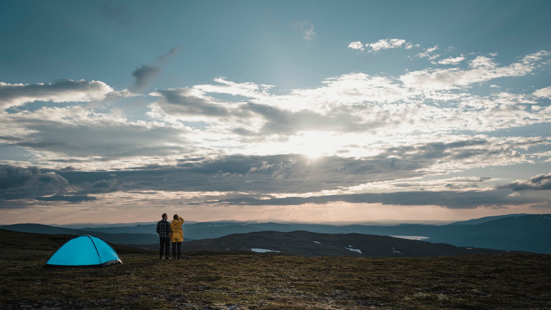 En man och en kvinna står på ett fjäll i solnedgången. I bilden syns också ett blått tält.