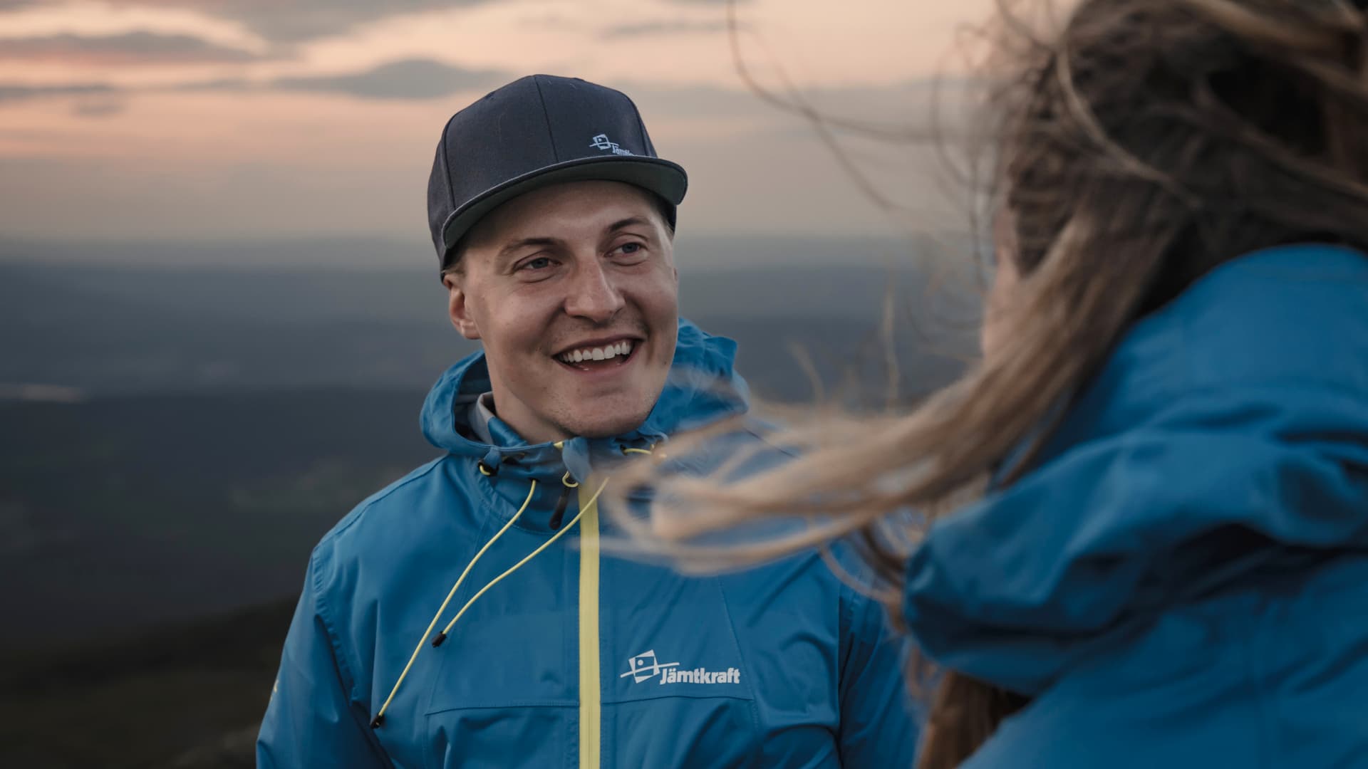 En ung man står på ett fjäll i skymningen och ler åt en kvinna som har ansiktet vänt från kameran.