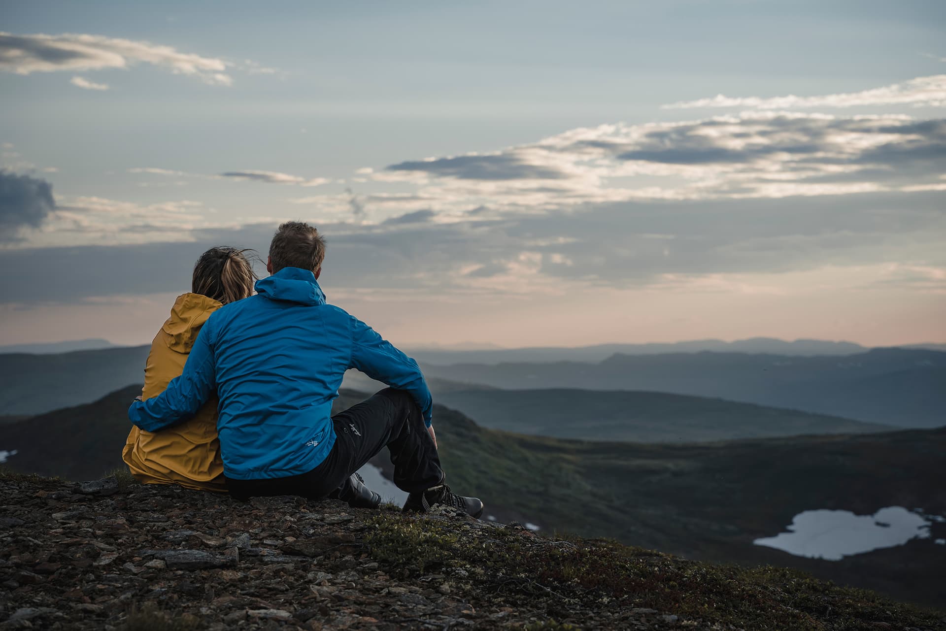 Ryggen på två personer som sitter på en fjäll och blickar ut över omgivningen.