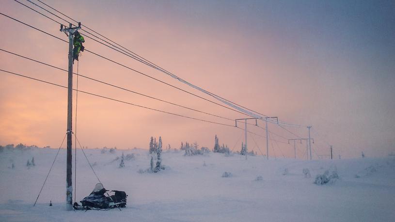 En medarbetare på Jämtkraft klättrar på en elnätsstolpe på ett fjäll i skymningen en vinterdag.