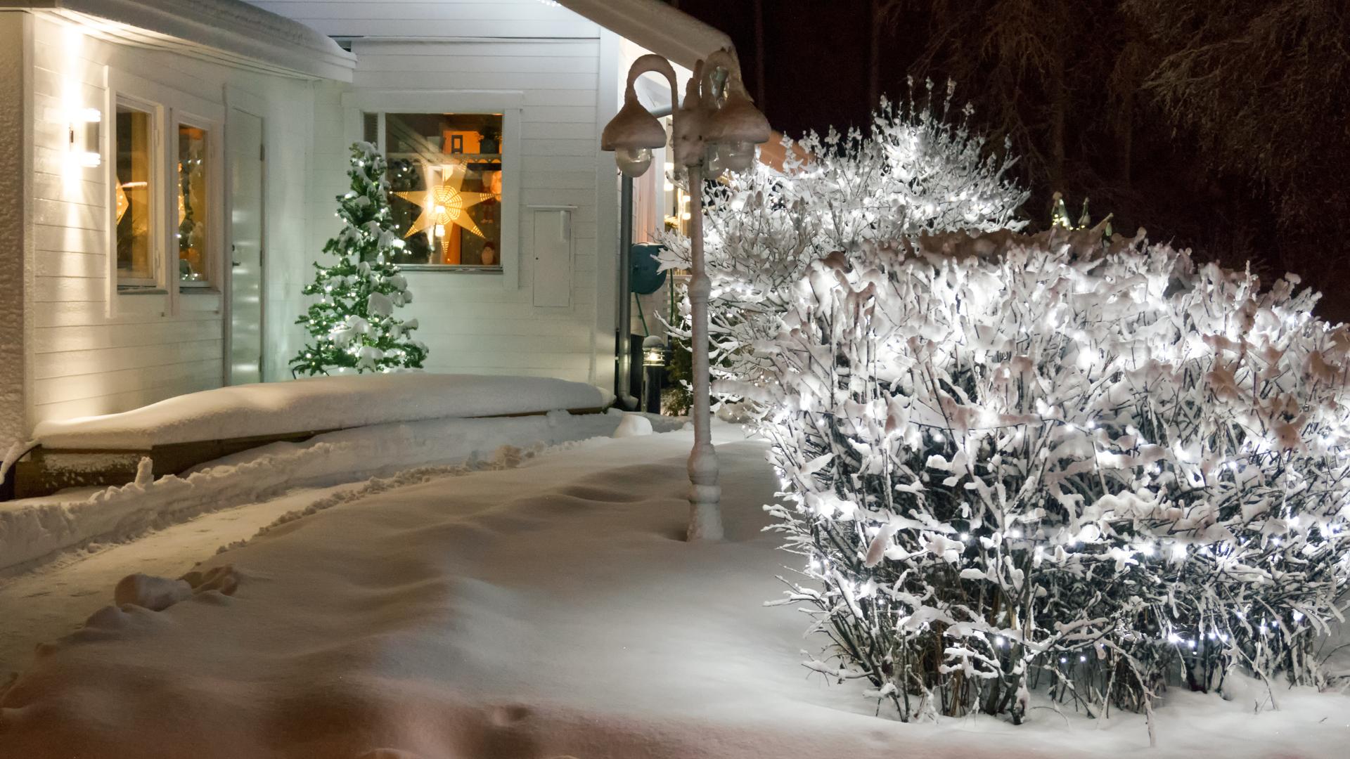En bild tagen utifrån på en del av ett vitt hus där man ser adventsstjärnor i fönstren. Utanför är det snö och på altanen står en adventsgran. Man skymtar även en upplyst trädgård.