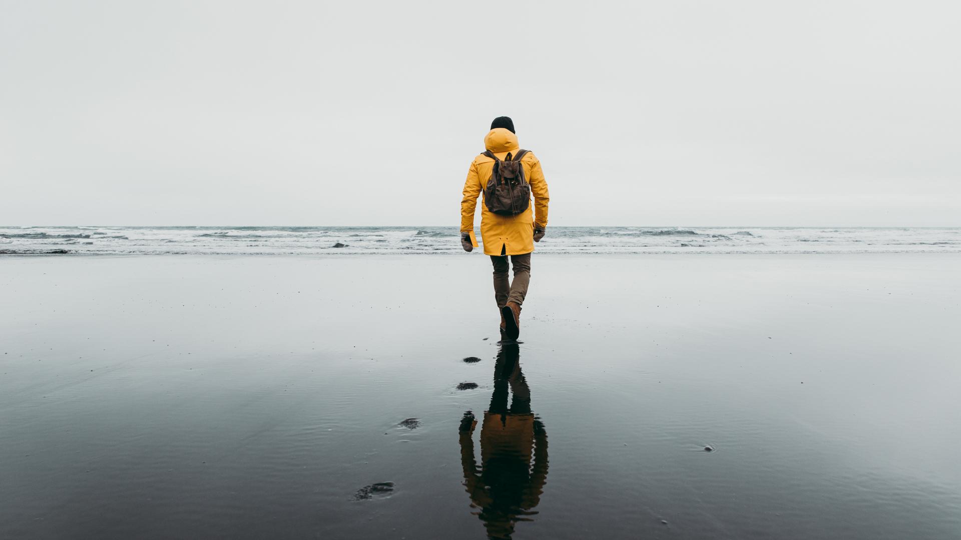Bild tagen bakifrån på en människa med gul jacka och ryggsäck som går på en strand en regnig dag.