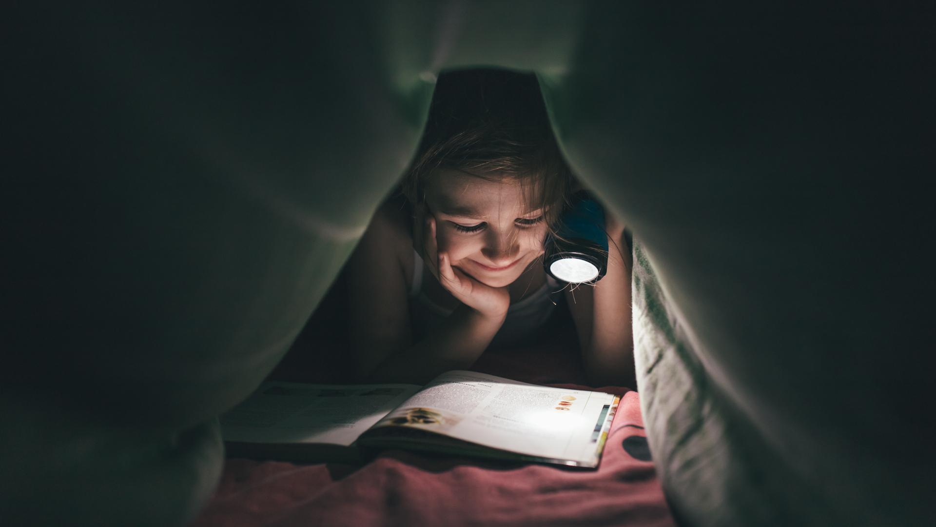 Ett barn läser en bok under en filt med en ficklampa i ett mörkt rum.