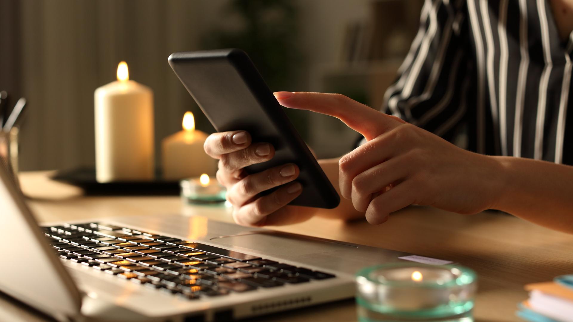 Närbild på en kvinnas händer som håller i en smartphone. Hon sitter vid en köksbord och på bordet syns en dator och några stearinljus.