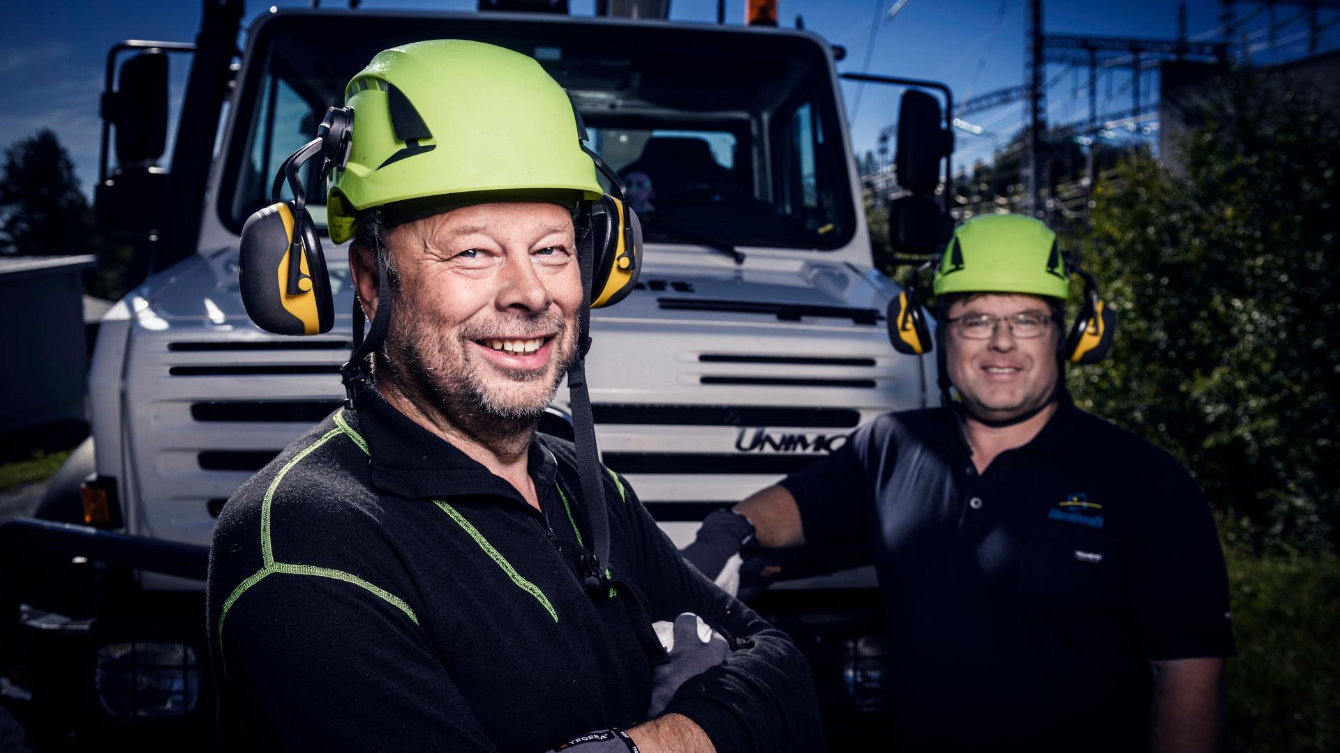 Porträtt av två män i arbetskläder och skyddshjälm som står framför en lastbil.