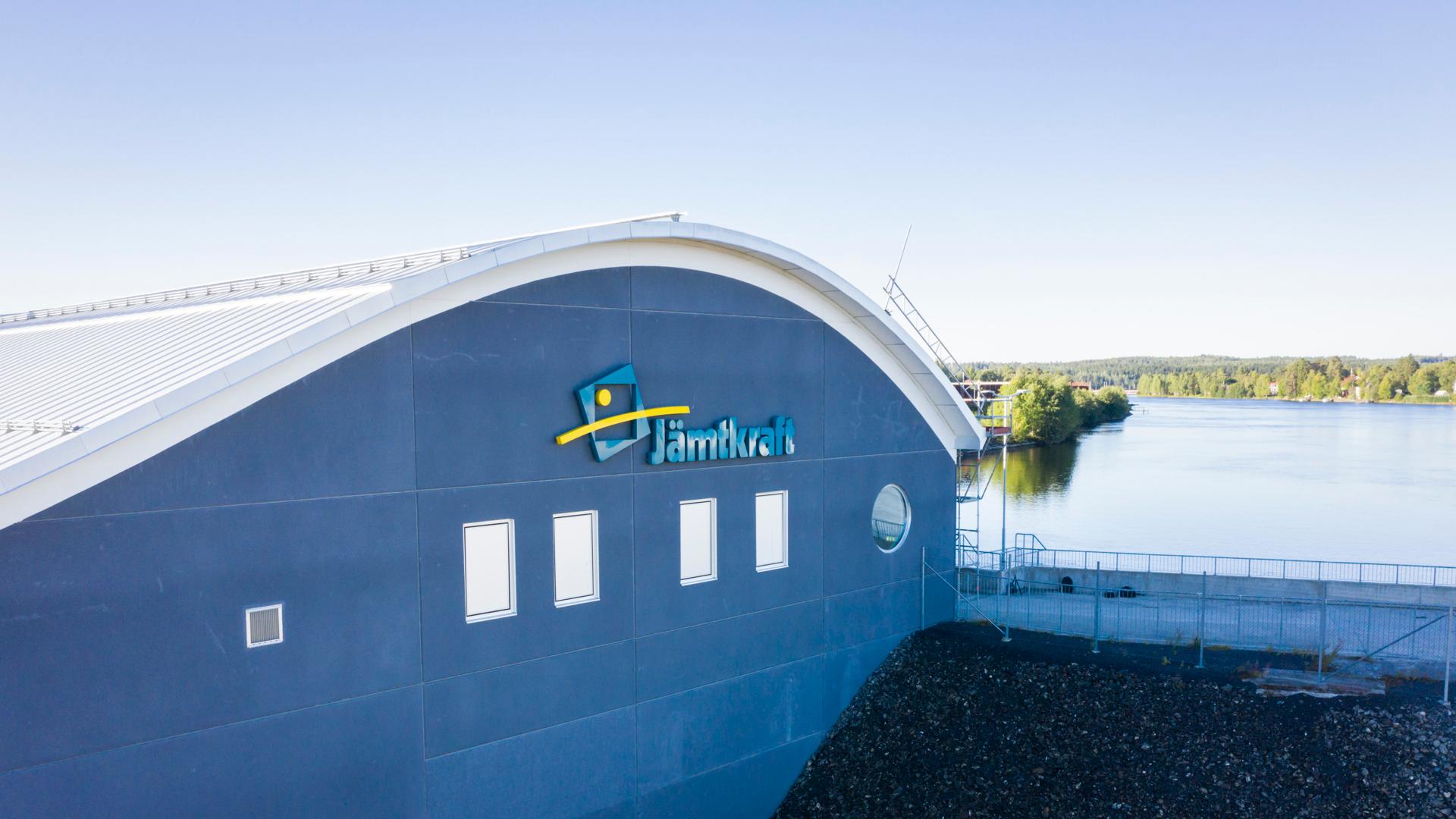 Sidan av vattenkraftverket i Hissmofors. På väggen sitter Jämtkrafts logotyp och i bakgrunden syns vatten vid kraftverket.