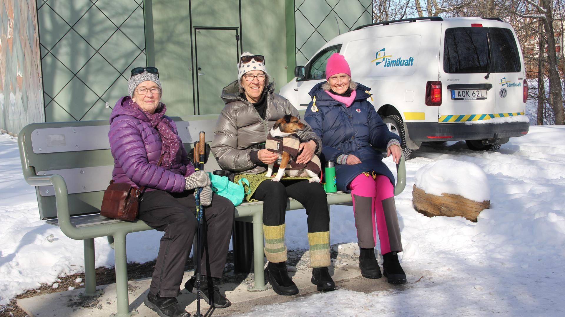 Tre kvinnor och en hund sitter på en bänk i vintrig miljö.