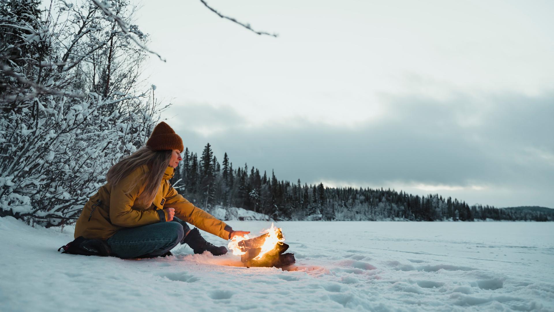 En kvinna i jacka och mössa sitter och värmer händerna över en öppen eld vid en sjö vintertid.