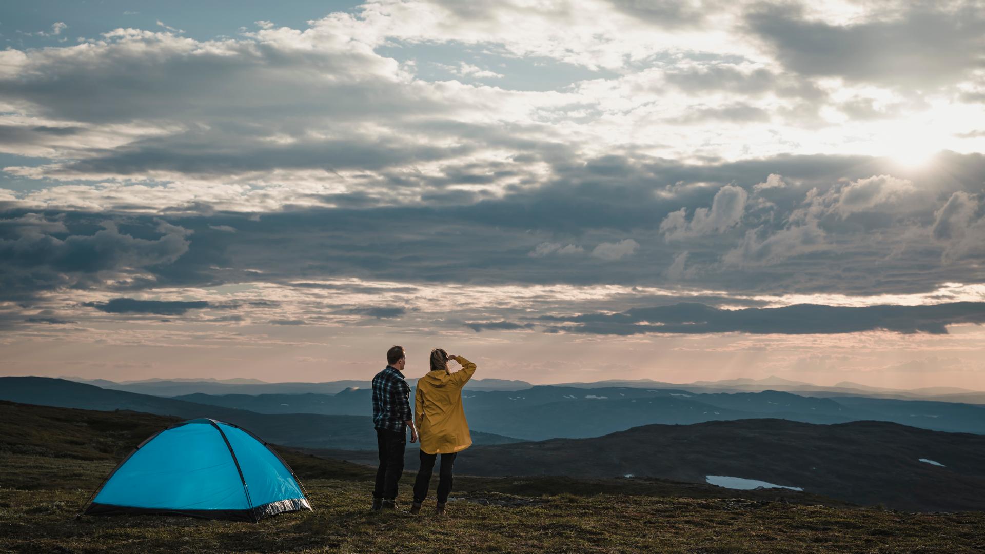 Två personer står med ryggen till på ett fjäll med utsikt över fjäll och sjö. Till vänster syns ett blått tält.