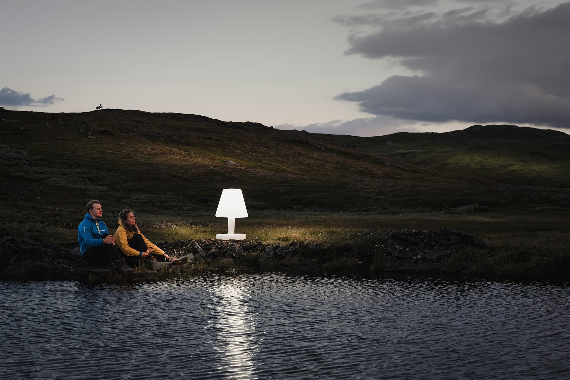 Två personer sitter på ett fjäll vid en vattendrag. Bredvid står en lysande lampa.