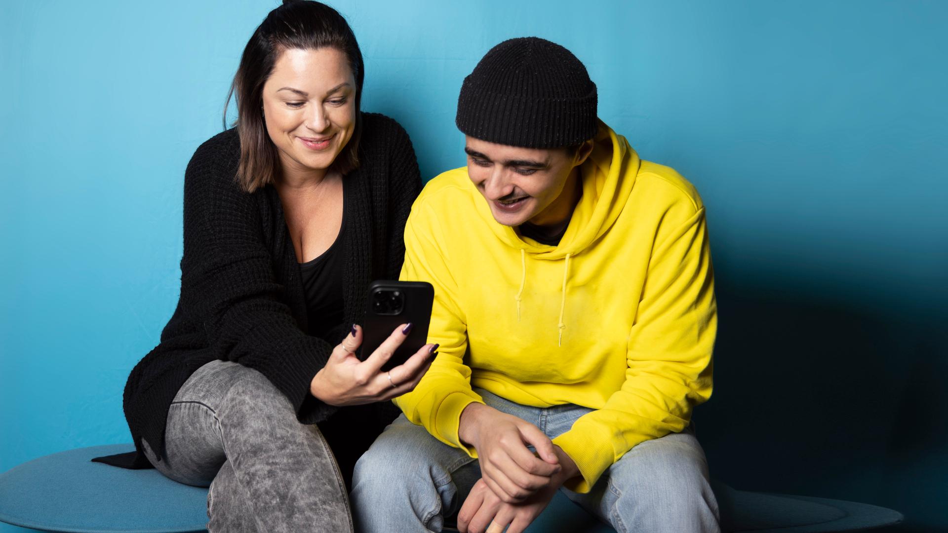 En ung man i gul tröja och svart mössa sitter bredvid en ung kvinna i svart tröja. De ler och tittar tillsammans på en mobiltelefon.