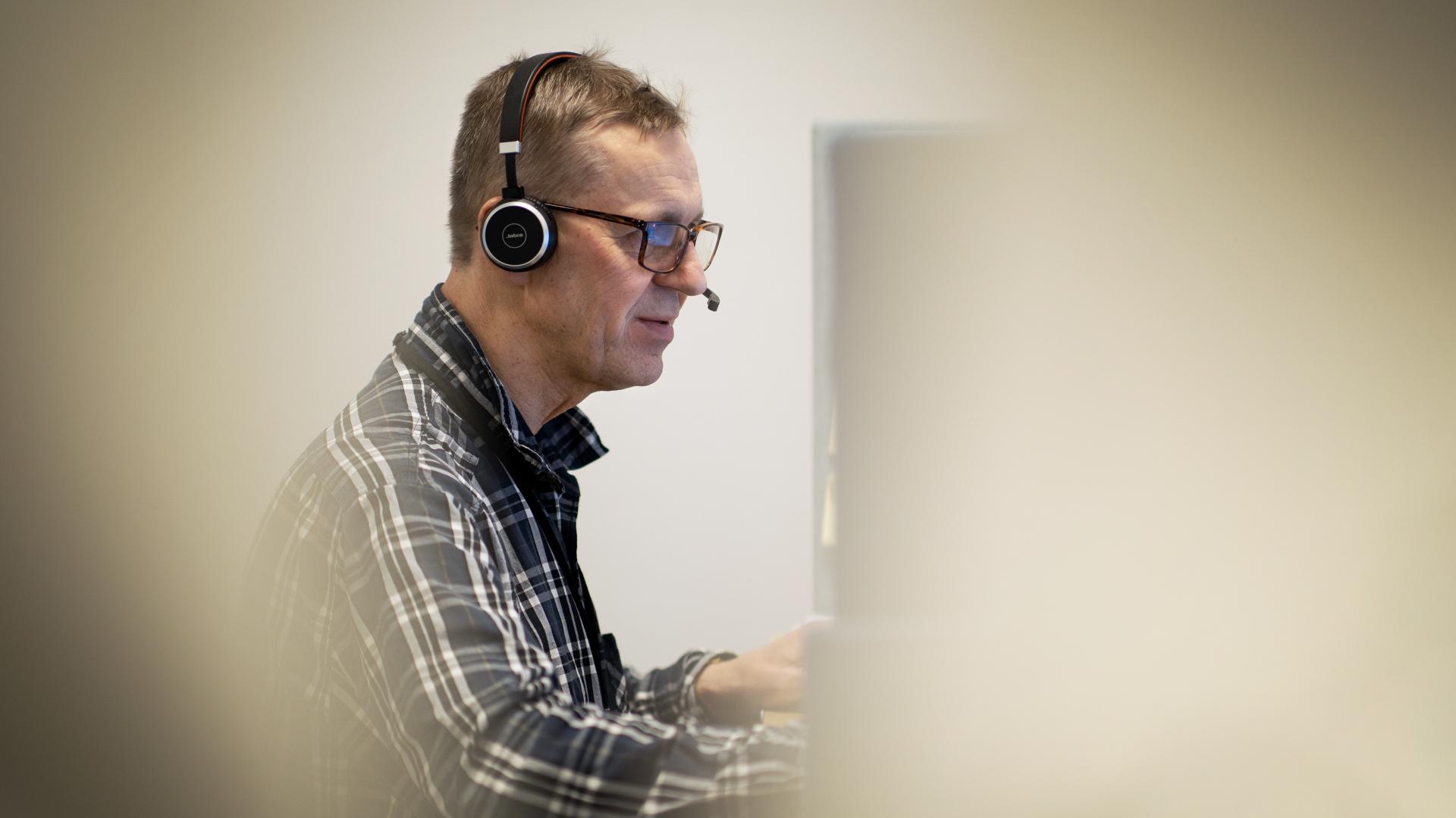 En man i rutig skjorta sitter med headset och tittar på en datorskärm.